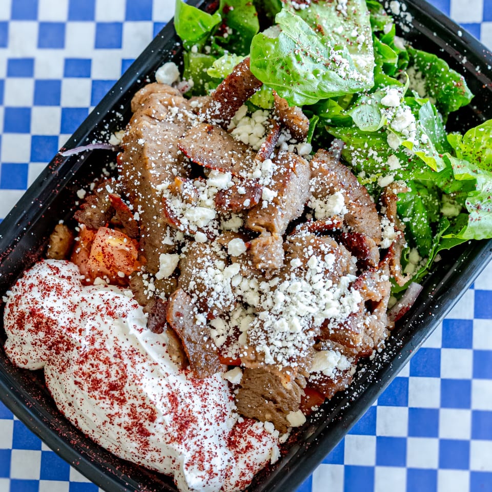 Grapeleaf Mediterranean Grille's gyro meat, feta, and salad in a black to-go container on a blue checkered surface.