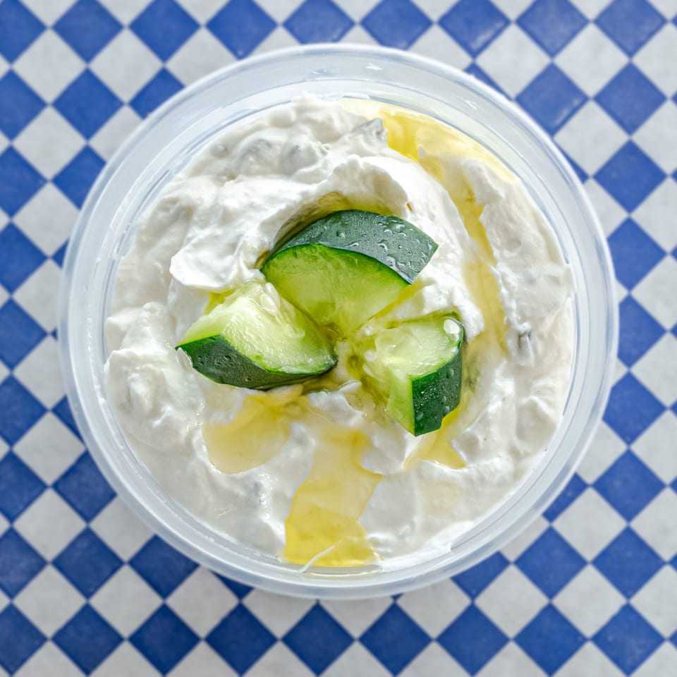 Tzatziki dip in a clear container, topped with cucumber slices and olive oil, on a blue and white checkered surface.